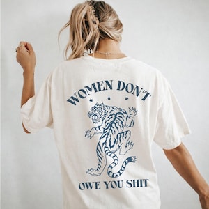 Pode incluir: Camiseta creme com um gráfico de tigre azul marinho e o texto "WOMEN DON'T OWE YOU SHIT." O tigre é retratado em um estilo ilustrativo. A camiseta tem mangas curtas e um corte folgado.