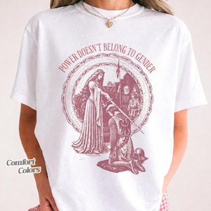 Può includere: T-shirt bianca con grafica bordeaux. La grafica presenta un'illustrazione in stile medievale all'interno di una cornice circolare, con la scritta "POWER DOESN'T BELONG TO GENDER" sopra. La maglietta ha anche le parole "Comfort Colors" e "white".
