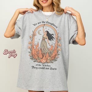 May include: A light grey t-shirt with the text "We are the Daughters of the Witches They could not Burn." The graphic features a woman in a dress, surrounded by flames and flowers. The shirt is a sports grey color.