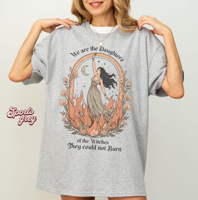 May include: A light grey t-shirt with the text "We are the Daughters of the Witches They could not Burn." The graphic features a woman in a dress, surrounded by flames and flowers. The shirt is a sports grey color.