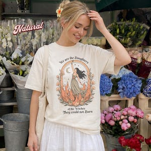 May include: Cream-colored t-shirt with a graphic of a woman in a flowing dress, surrounded by flames and floral elements. The text on the shirt reads "We are the Daughters of the Witches They could not Burn."