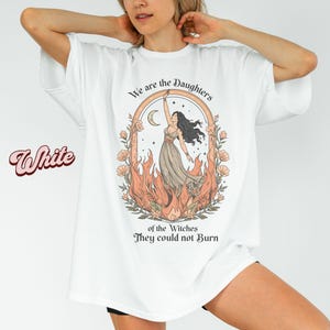 May include: White t-shirt with a graphic of a woman in a dress, surrounded by flames and flowers, with the text "We are the Daughters of the Witches They could not Burn". The word "White" is written in a cursive font.