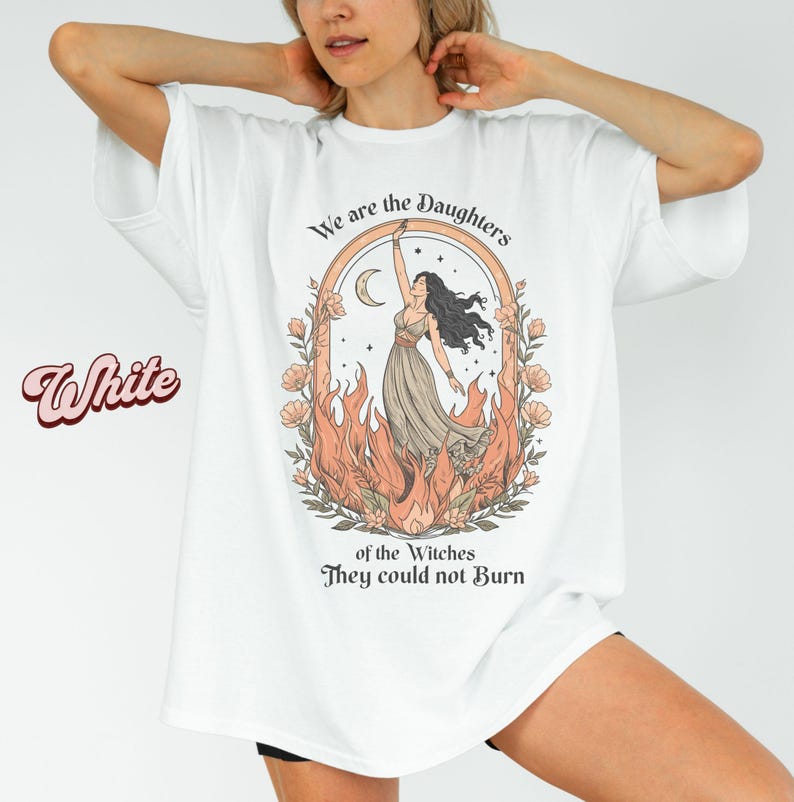 May include: White t-shirt with a graphic of a woman in a dress, surrounded by flames and flowers, with the text "We are the Daughters of the Witches They could not Burn". The word "White" is written in a cursive font.