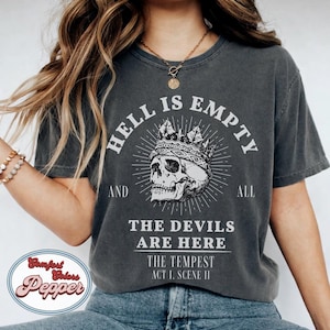 Op de afbeelding: Donkergrijs t-shirt met de tekst "HELL IS EMPTY AND ALL THE DEVILS ARE HERE" en een schedel met een kroon. De tekst "THE TEMPEST ACT 1. SCENE II" staat ook op de shirt. Het shirt heeft een vintage, distressed look.