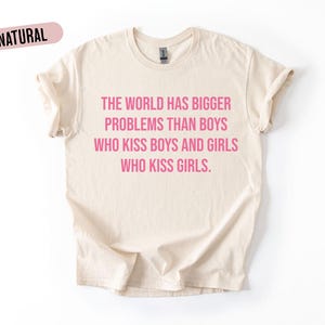 May include: A cream-coloured t-shirt with the word "NATURAL" in a pink oval at the top left. The shirt features the text "THE WORLD HAS BIGGER PROBLEMS THAN BOYS WHO KISS BOYS AND GIRLS WHO KISS GIRLS" in pink.