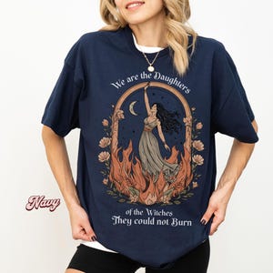 May include: Navy blue t-shirt featuring a graphic of a woman in a flowing dress, framed by flames and flowers, with the text "We are the Daughters of the Witches They could not Burn."