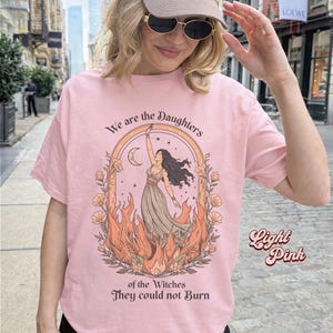 May include: Light pink t-shirt with the text "We are the Daughters of the Witches They could not Burn." The graphic features a woman in a dress, surrounded by flames, with a crescent moon above her. The words "Light Pink" are written on the right side.