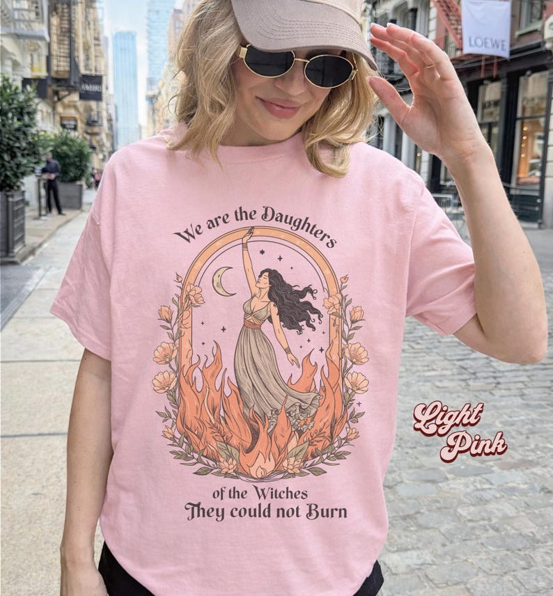 May include: Light pink t-shirt with the text "We are the Daughters of the Witches They could not Burn." The graphic features a woman in a dress, surrounded by flames, with a crescent moon above her. The words "Light Pink" are written on the right side.