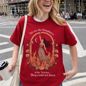 May include: Red t-shirt with a graphic design featuring a woman in a dress, surrounded by flames and flowers, with the text "We are the Daughters of the Witches They could not Burn."