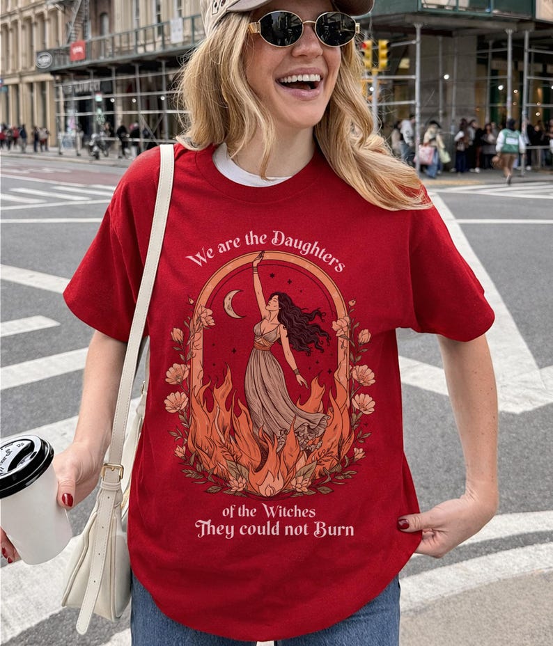 May include: Red t-shirt with a graphic design featuring a woman in a dress, surrounded by flames and flowers, with the text "We are the Daughters of the Witches They could not Burn."
