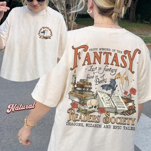 Op de afbeelding: Crèmekleurig T-shirt met een afbeelding op de voor- en achterkant. De afbeelding op de achterkant luidt "Proud Member of the Fantasy Readers Society" met illustraties van boeken, draken en rozen. De afbeelding op de voorkant heeft de tekst "Fantasy Readers Society".