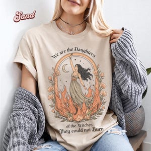 May include: Sand-colored t-shirt featuring a graphic of a woman in a flowing dress, framed by flames and flowers, with the text "We are the Daughters of the Witches They could not Burn". The design includes a crescent moon and a rainbow arch.