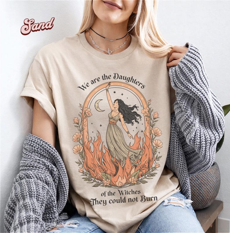 May include: Sand-colored t-shirt featuring a graphic of a woman in a flowing dress, framed by flames and flowers, with the text "We are the Daughters of the Witches They could not Burn". The design includes a crescent moon and a rainbow arch.