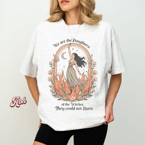 May include: A light gray t-shirt featuring a graphic of a woman in a flowing dress, surrounded by flames and flowers. The text "We are the Daughters of the Witches They could not Burn" is arched above and below the illustration.