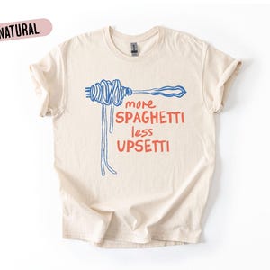 Puede incluir: Camiseta color crema con el texto "more spaghetti less upsetti" en rojo y azul. El diseño presenta un tenedor con espaguetis y fideos colgantes. La palabra "NATURAL" está en un óvalo rosa.