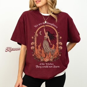 May include: Maroon t-shirt with a white crew neck featuring a graphic of a woman in a dress with flames and the text "We are the Daughters of the Witches They could not Burn".