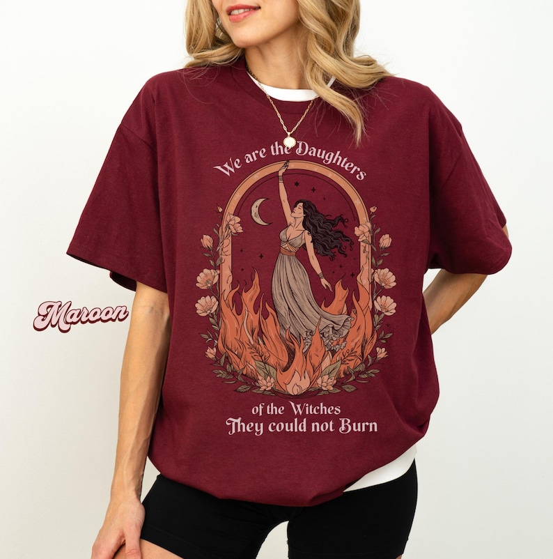 May include: Maroon t-shirt with a white crew neck featuring a graphic of a woman in a dress with flames and the text "We are the Daughters of the Witches They could not Burn".