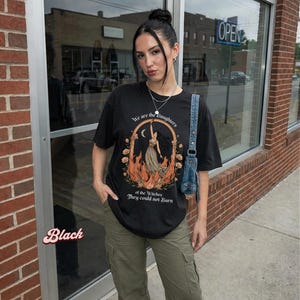 May include: Black t-shirt with the text "We are the Daughters of the Witches They could not Burn." The graphic features a woman in a dress with flames and a crescent moon. The shirt is paired with olive green cargo pants.