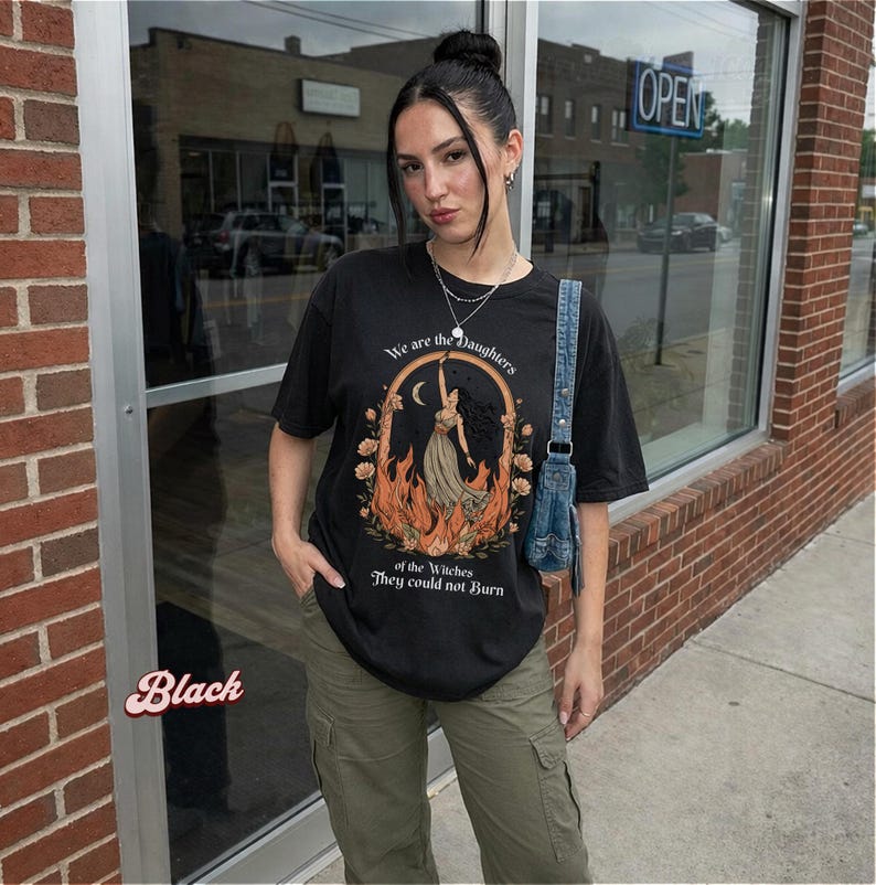 May include: Black t-shirt with the text "We are the Daughters of the Witches They could not Burn." The graphic features a woman in a dress with flames and a crescent moon. The shirt is paired with olive green cargo pants.
