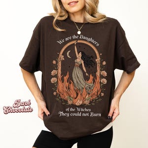 May include: Dark chocolate brown t-shirt with a graphic design. The design features a woman in a flowing dress, surrounded by flames, with the text "We are the Daughters of the Witches They could not Burn."
