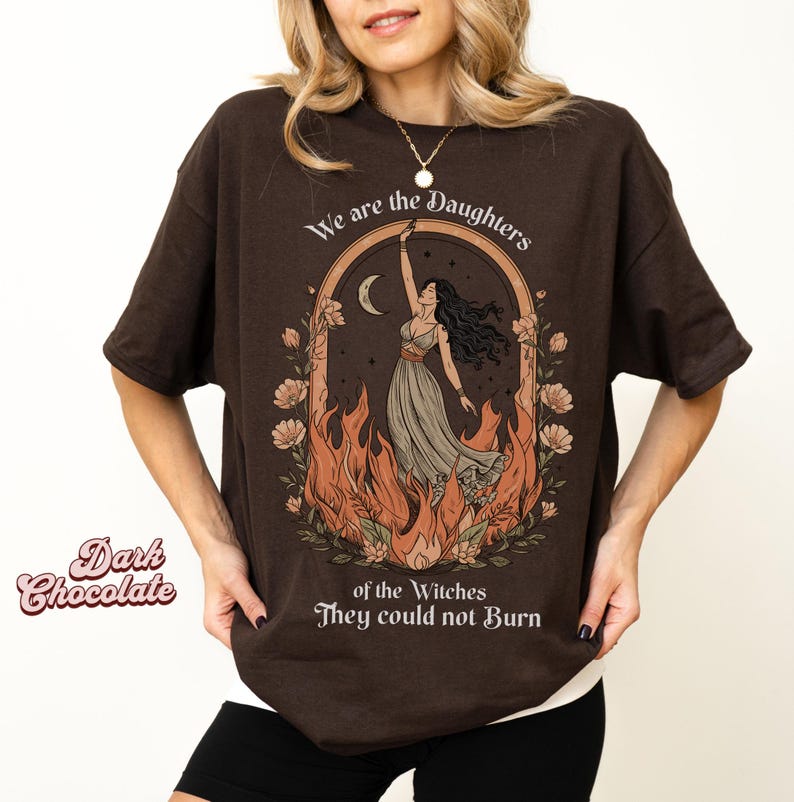 May include: Dark chocolate brown t-shirt with a graphic design. The design features a woman in a flowing dress, surrounded by flames, with the text "We are the Daughters of the Witches They could not Burn."