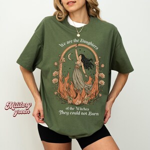May include: Military green t-shirt with a graphic of a woman in a dress reaching towards a crescent moon, surrounded by flames and flowers. Text reads "We are the Daughters of the Witches They could not Burn."