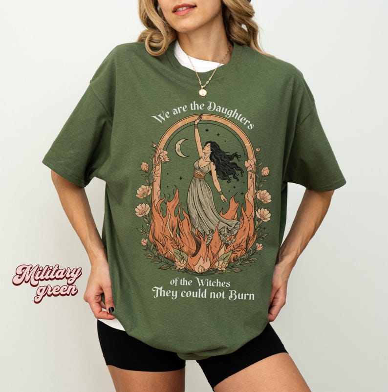 May include: Military green t-shirt with a graphic of a woman in a dress reaching towards a crescent moon, surrounded by flames and flowers. Text reads "We are the Daughters of the Witches They could not Burn."