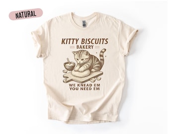 Può includere: T-shirt color crema con la scritta "Kitty Biscuits Bakery" e una grafica di un gatto che impasta. Il testo "We Knead Em You Need Em" è sotto. La maglietta ha maniche corte e girocollo.