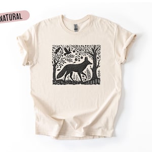 Puede incluir: Una camiseta color crema con un gráfico negro de un zorro en una escena de bosque con árboles y pájaros. La palabra "NATURAL" está impresa en un óvalo rosa en la esquina superior izquierda. La camiseta tiene mangas cortas.