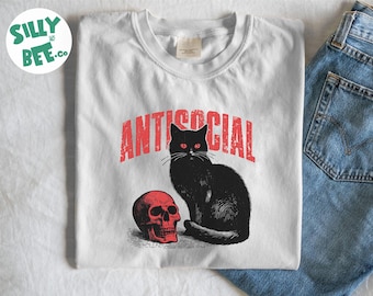 Könnte beinhalten: Ein weißes T-Shirt mit Rundhalsausschnitt und einem Grafikdruck einer schwarzen Katze, die neben einem roten menschlichen Schädel sitzt. Über der Katze ist das Wort ANTISOCIAL in einer abgenutzten roten Schrift gedruckt. Das T-Shirt liegt neben einer blauen Jeans.