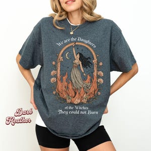 May include: Dark heather gray t-shirt with a graphic featuring a woman in a flowing dress, framed by flames and flowers, with the text "We are the Daughters of the Witches They could not Burn."