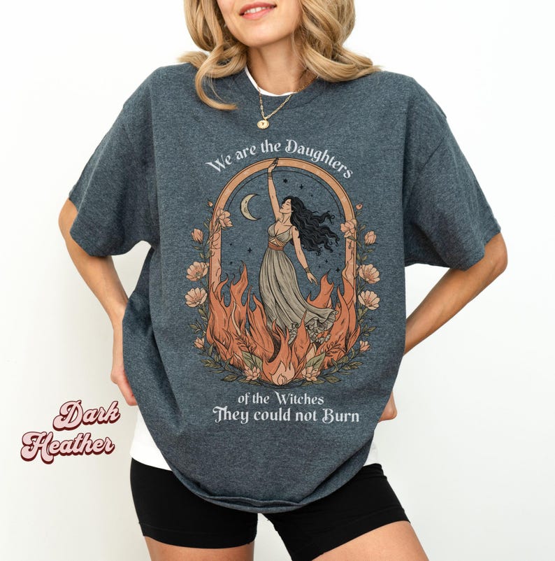 May include: Dark heather gray t-shirt with a graphic featuring a woman in a flowing dress, framed by flames and flowers, with the text "We are the Daughters of the Witches They could not Burn."