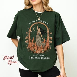 May include: Forest green t-shirt with a graphic of a woman in a dress reaching towards a crescent moon, surrounded by flames and flowers. Text reads "We are the Daughters of the Witches They could not Burn."