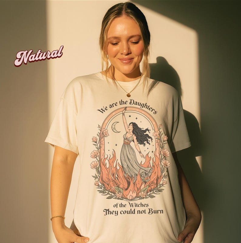 May include: Cream-colored t-shirt featuring a graphic of a woman with long black hair, reaching towards a crescent moon. The design is framed by flames and flowers, with the text "We are the Daughters of the Witches They could not Burn."