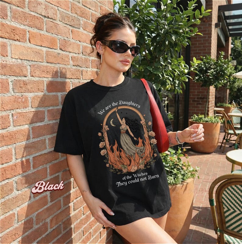 May include: Black t-shirt with a graphic of a woman in a flowing dress, framed by flames and flowers, with the text "We are the Daughters of the Witches They could not Burn". The word "Black" is printed in a pink script font.