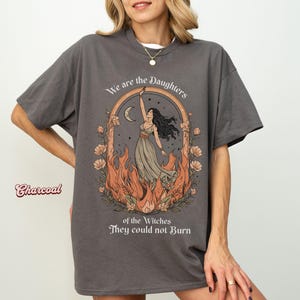 May include: Charcoal-colored t-shirt with a graphic featuring a woman in a flowing dress, framed by a floral arch, flames, and the text "We are the Daughters of the Witches They could not Burn."