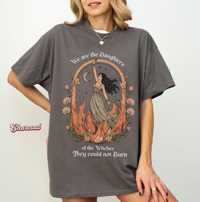 May include: Charcoal-colored t-shirt with a graphic featuring a woman in a flowing dress, framed by a floral arch, flames, and the text "We are the Daughters of the Witches They could not Burn."