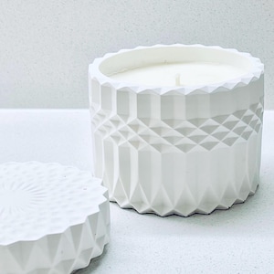 May include: A white candle in a geometric patterned container with a matching lid. The candle has a single wick and is set on a white surface. The container has a faceted design and a textured lid, creating a modern aesthetic.