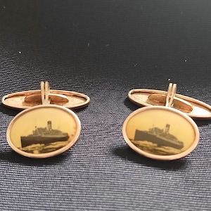 May include: A pair of gold-toned cufflinks. Each cufflink features an oval-shaped face with a sepia-toned image of a ship. The cufflinks have a classic design with a hinged backing for secure fastening.