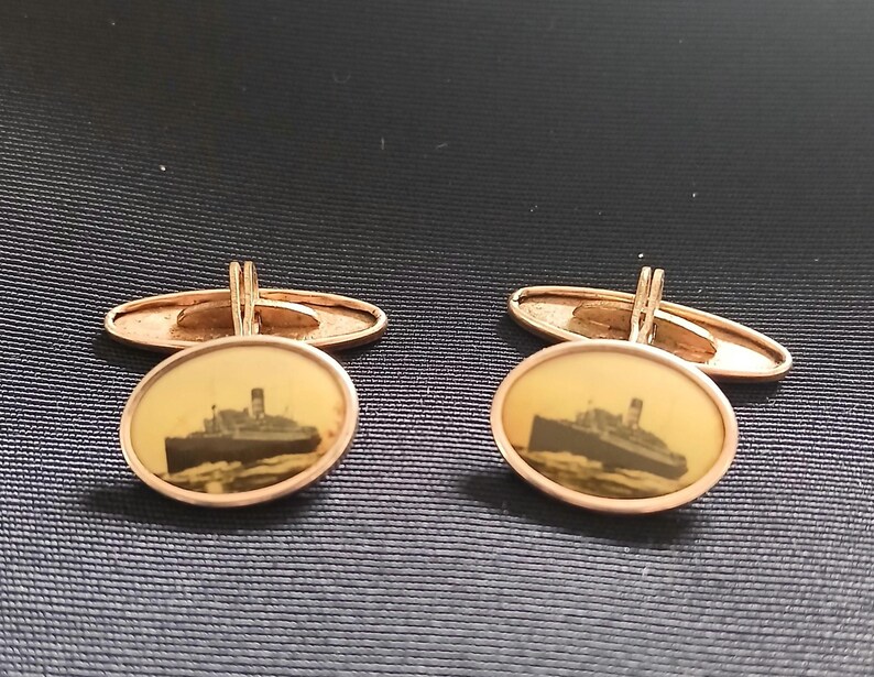 May include: A pair of gold-toned cufflinks. Each cufflink features an oval-shaped face with a sepia-toned image of a ship. The cufflinks have a classic design with a hinged backing for secure fastening.