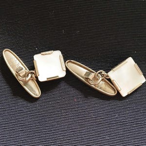 May include: A pair of gold-toned cufflinks with square, off-white insets. The cufflinks have a classic design with a chain connecting the two parts. The background is a dark blue fabric.