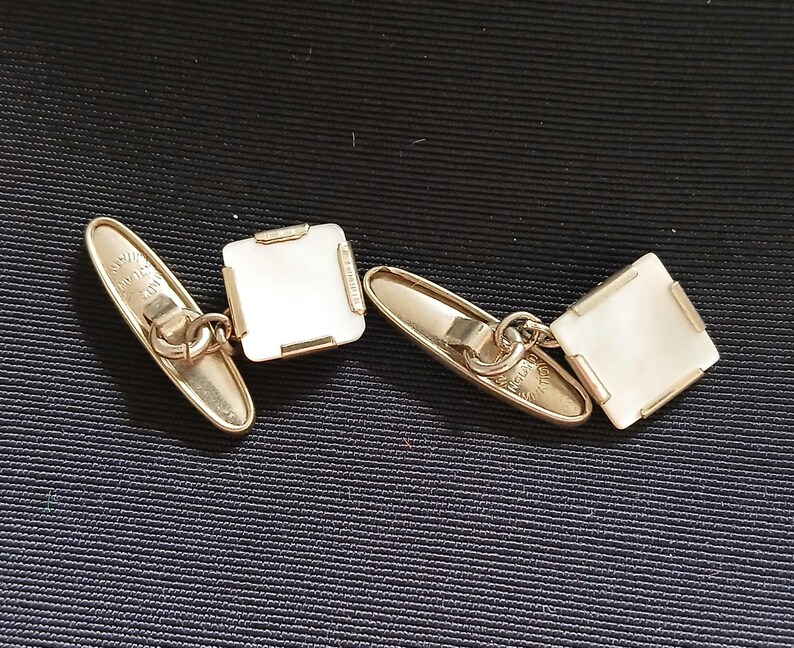 May include: A pair of gold-toned cufflinks with square, off-white insets. The cufflinks have a classic design with a chain connecting the two parts. The background is a dark blue fabric.
