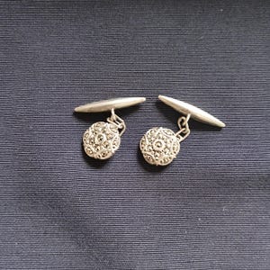 May include: A pair of silver cufflinks with a round, textured design. The cufflinks have a decorative, circular face with a detailed pattern. The background is a dark blue fabric.