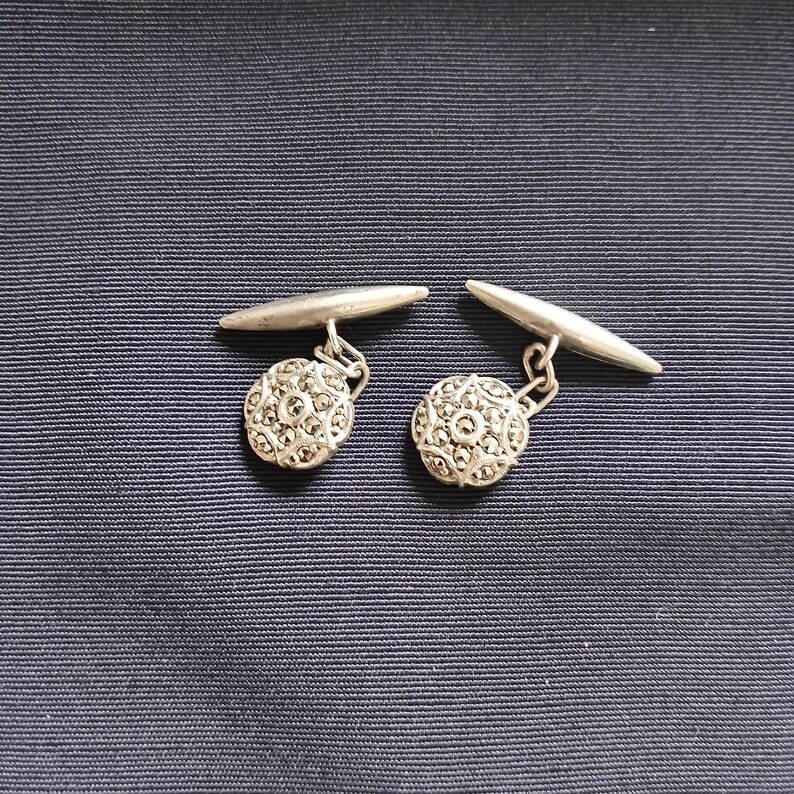 May include: A pair of silver cufflinks with a round, textured design. The cufflinks have a decorative, circular face with a detailed pattern. The background is a dark blue fabric.