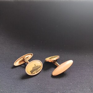 May include: A pair of gold-toned cufflinks. One cufflink features an oval design with a sepia-toned image of a ship. The other cufflink has a solid oval design. The cufflinks are displayed on a dark blue surface.