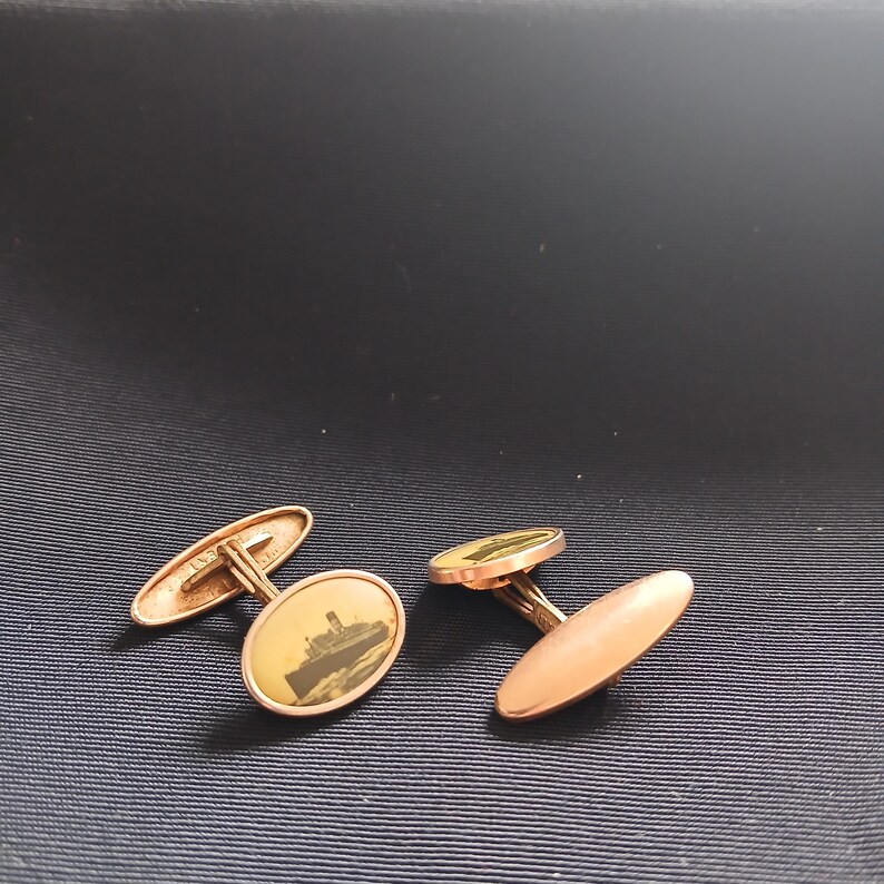 May include: A pair of gold-toned cufflinks. One cufflink features an oval design with a sepia-toned image of a ship. The other cufflink has a solid oval design. The cufflinks are displayed on a dark blue surface.