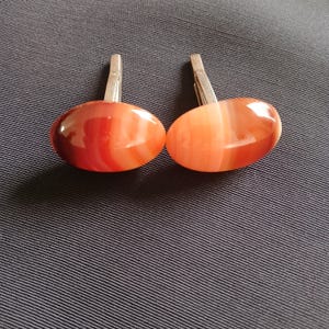 May include: A pair of cufflinks with oval-shaped, polished agate stones in shades of red and orange. The stones have a striped pattern and are set on silver-colored metal clasps. The cufflinks are displayed on a dark blue textured surface.