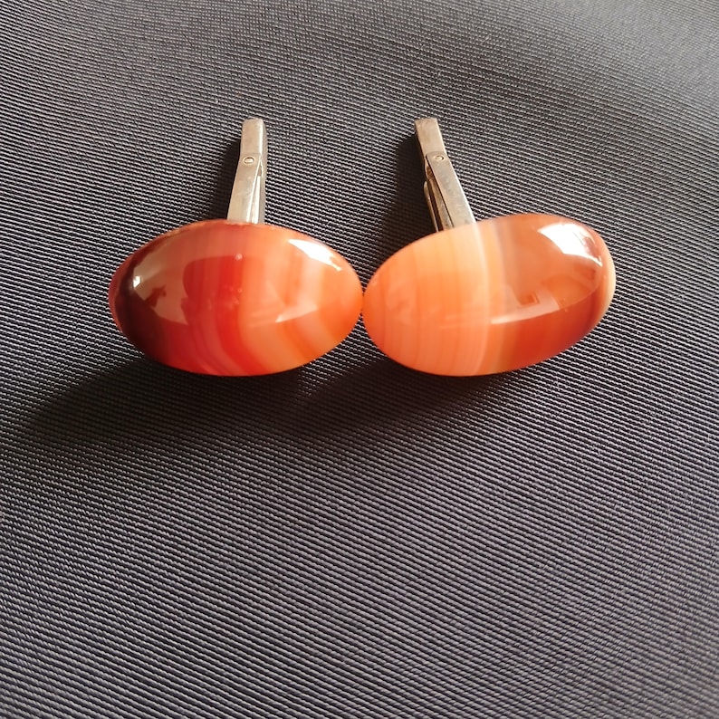 May include: A pair of cufflinks with oval-shaped, polished agate stones in shades of red and orange. The stones have a striped pattern and are set on silver-colored metal clasps. The cufflinks are displayed on a dark blue textured surface.