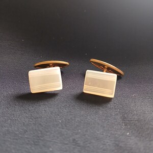 May include: A pair of vintage cufflinks. Each cufflink features a rectangular, off-white face with three horizontal lines and a gold-colored backing. The cufflinks are displayed on a dark blue surface.