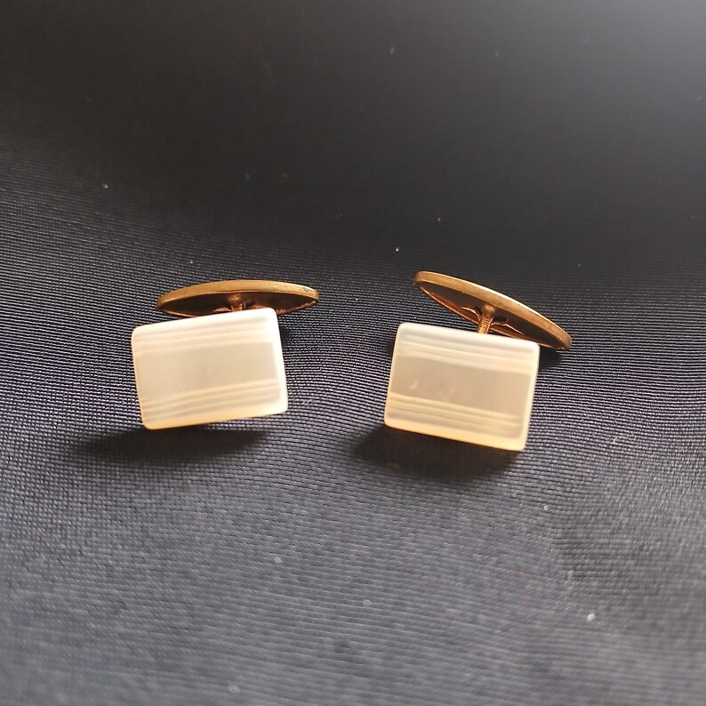 May include: A pair of vintage cufflinks. Each cufflink features a rectangular, off-white face with three horizontal lines and a gold-colored backing. The cufflinks are displayed on a dark blue surface.